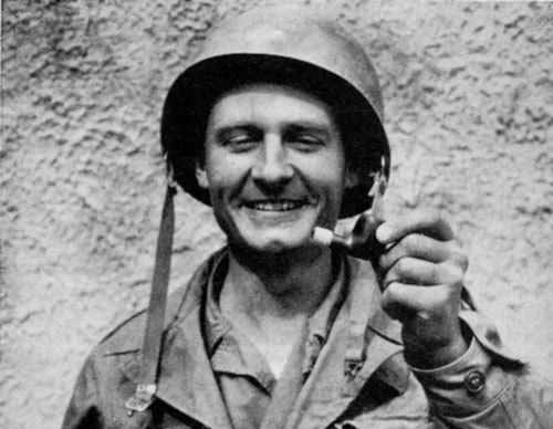 Chaplain Emil Kapaun smiles as he holds up a pipe broken by a sniper’s bullet.